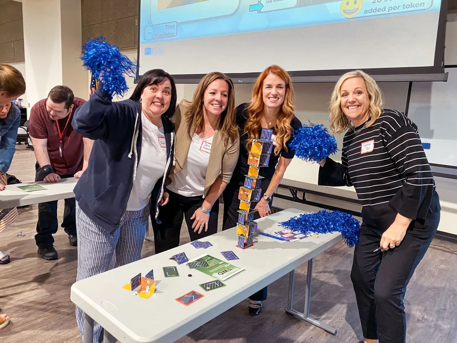 Four educators celebrate with pom-poms beside the card tower their team built, a ProSolve timer on screen behind them